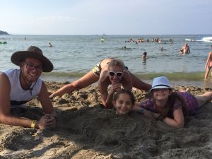 Z кемп, ден 16 - на плажа, дете заровено до главата в пясъка и други около него, морето е на заден план
